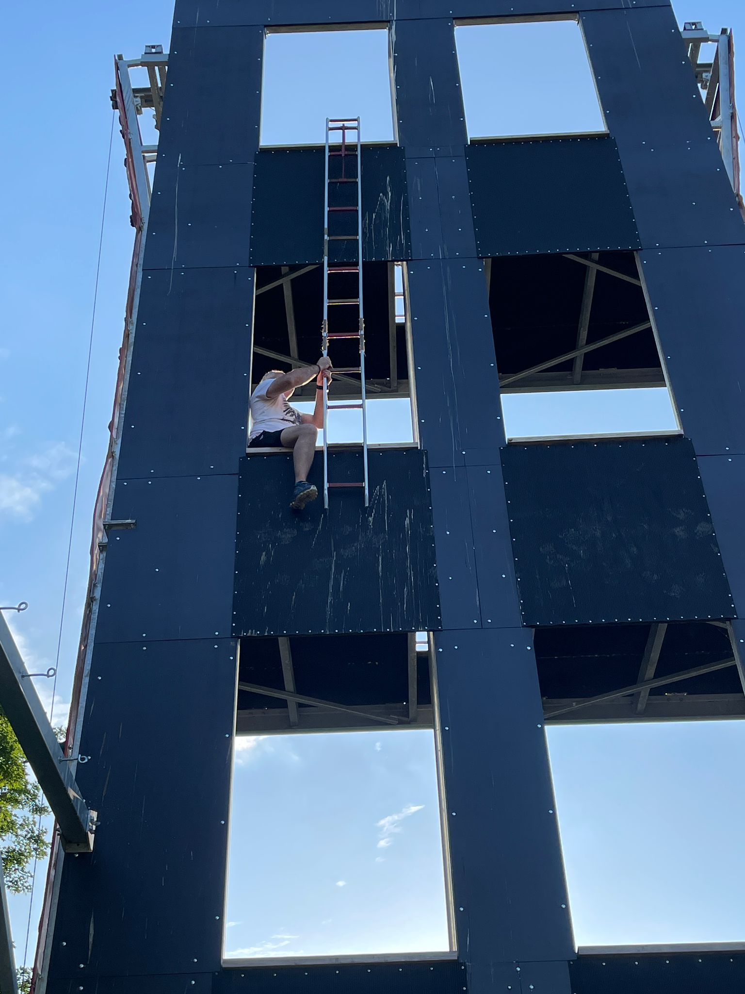 Instruktor z parapetu pierwszego piętra wieży strażackiej wysuwa drabinę, aby zaczepić ją o parapet drugiego piętra.