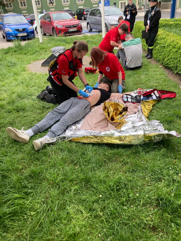 Na pierwszym planie- ratownicy udzielają pomocy poszkodwanemu z raną klatki piersiowej, w tle - drugi poszkodowany otrzymuje pomoc psychologiczną