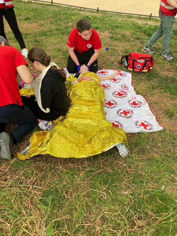 Poszkodowany owinięty folią termoizolacyjną leży na kocu. Ratownik stabilizuje głowę poszkodowanego.